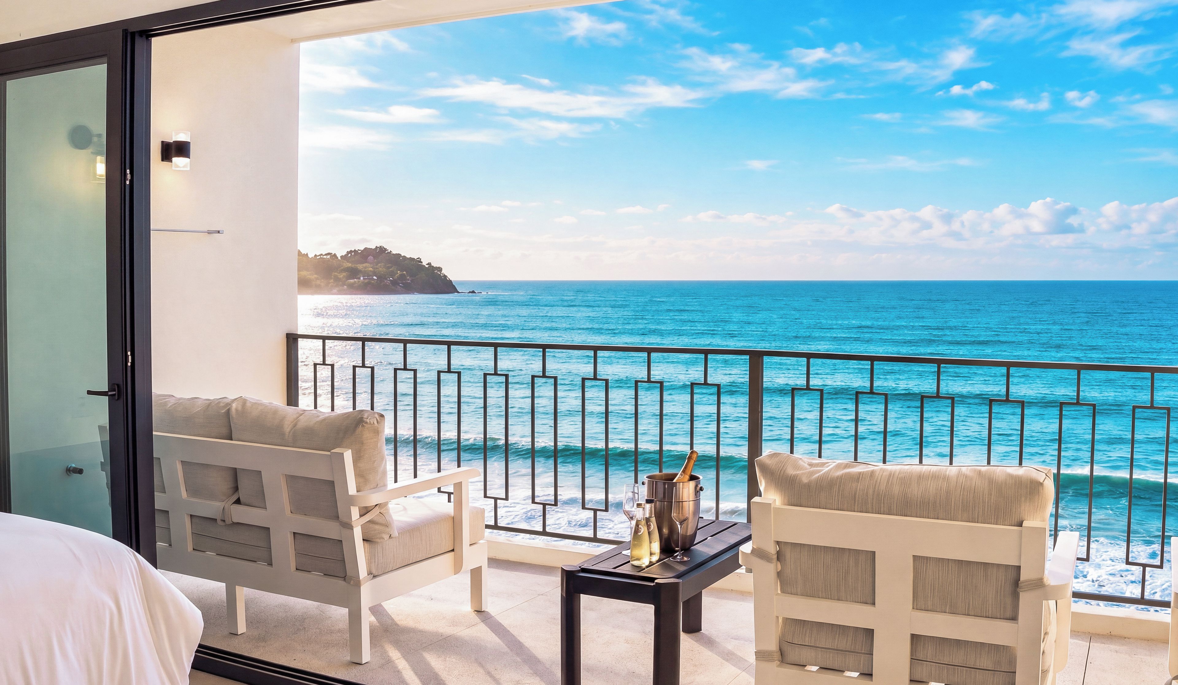A balcony with a view of the sea, two white chairs, a small table with a bottle and glass, and a bed with white bedding.
