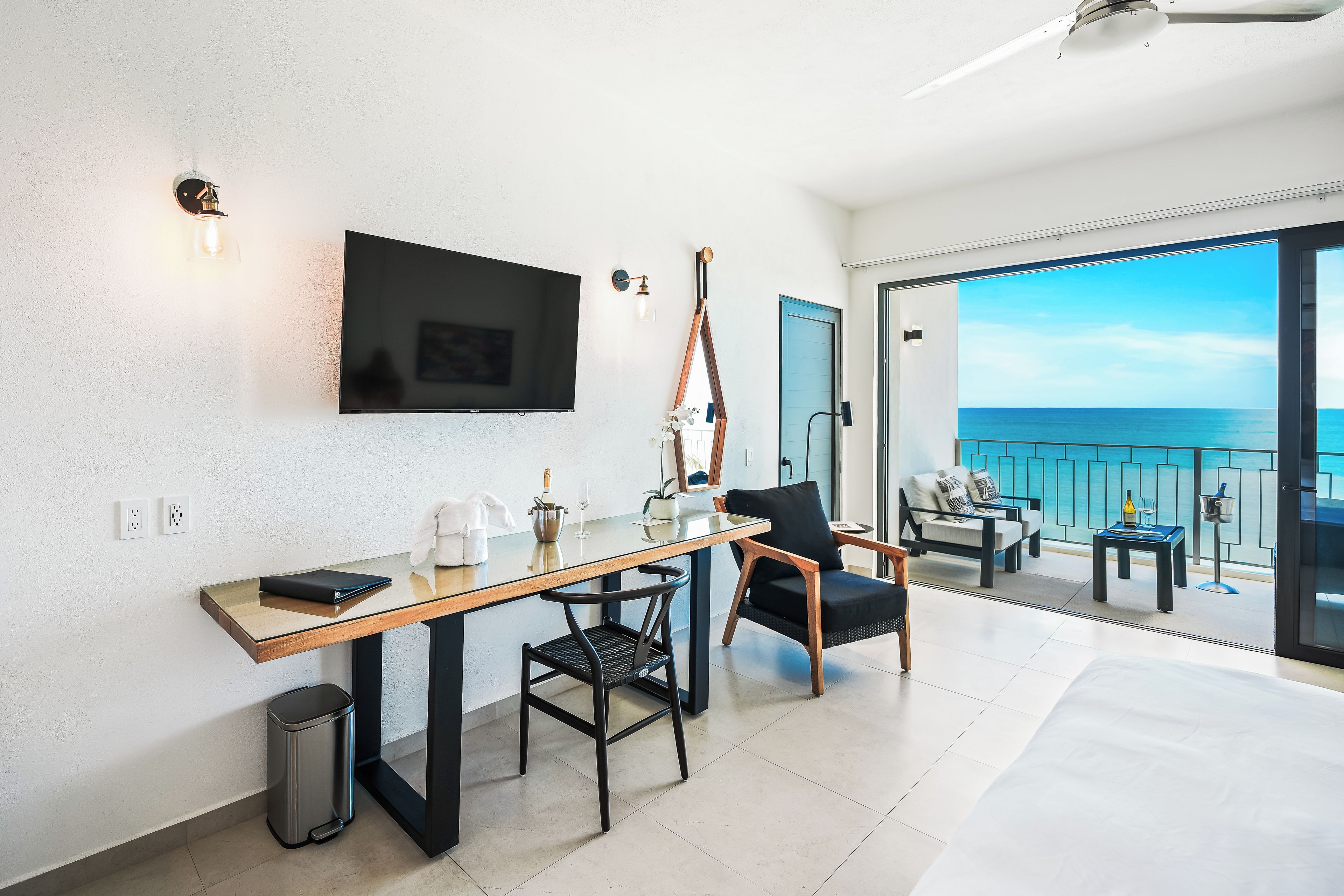 Habitación de hotel moderna con televisor de pantalla plana, escritorio de madera, una silla, un cesto de basura pequeño y balcón con vista al mar.