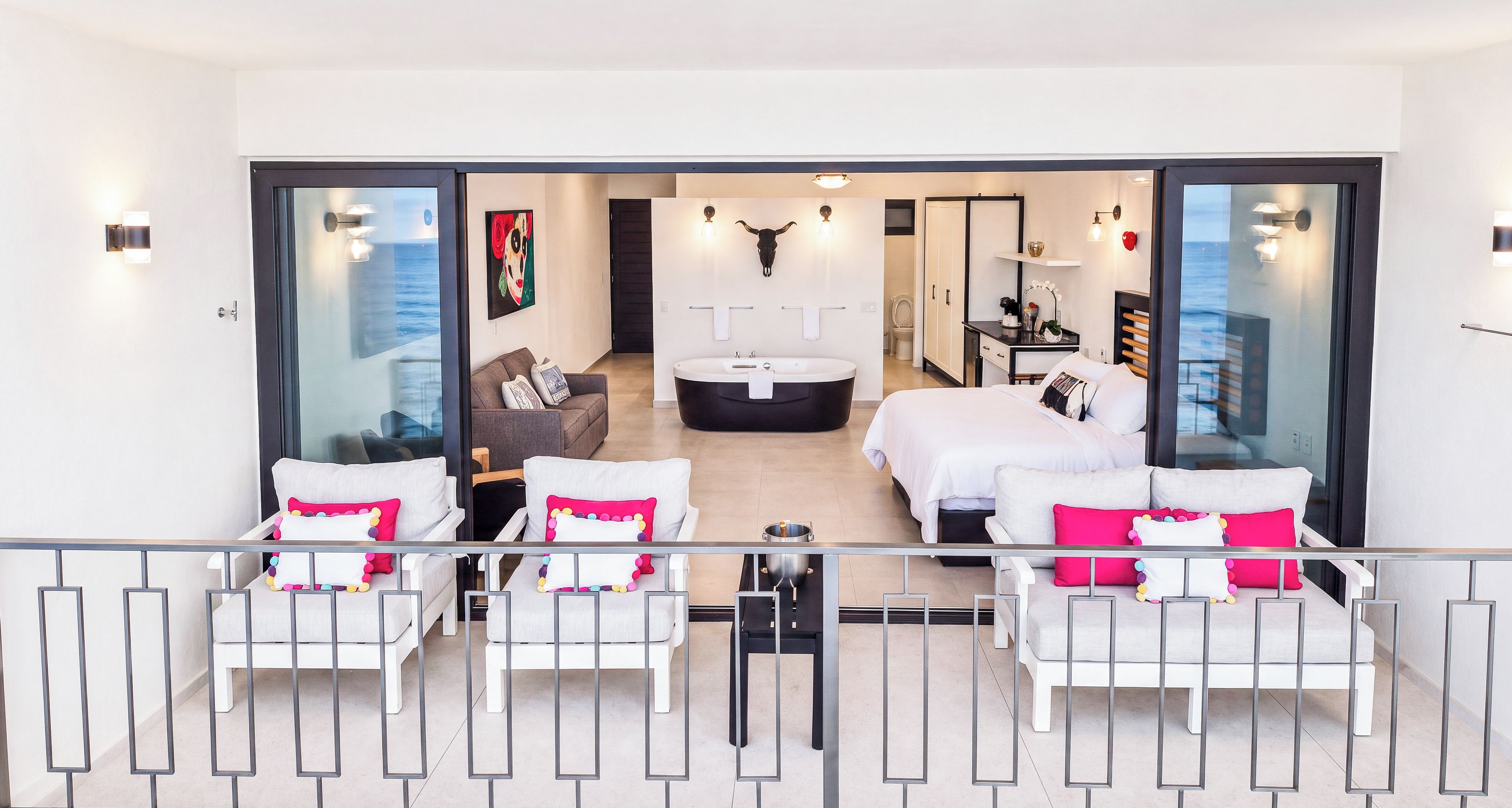 A modern interior with a living area, a large bed, a bathtub, and a view through a glass door.
