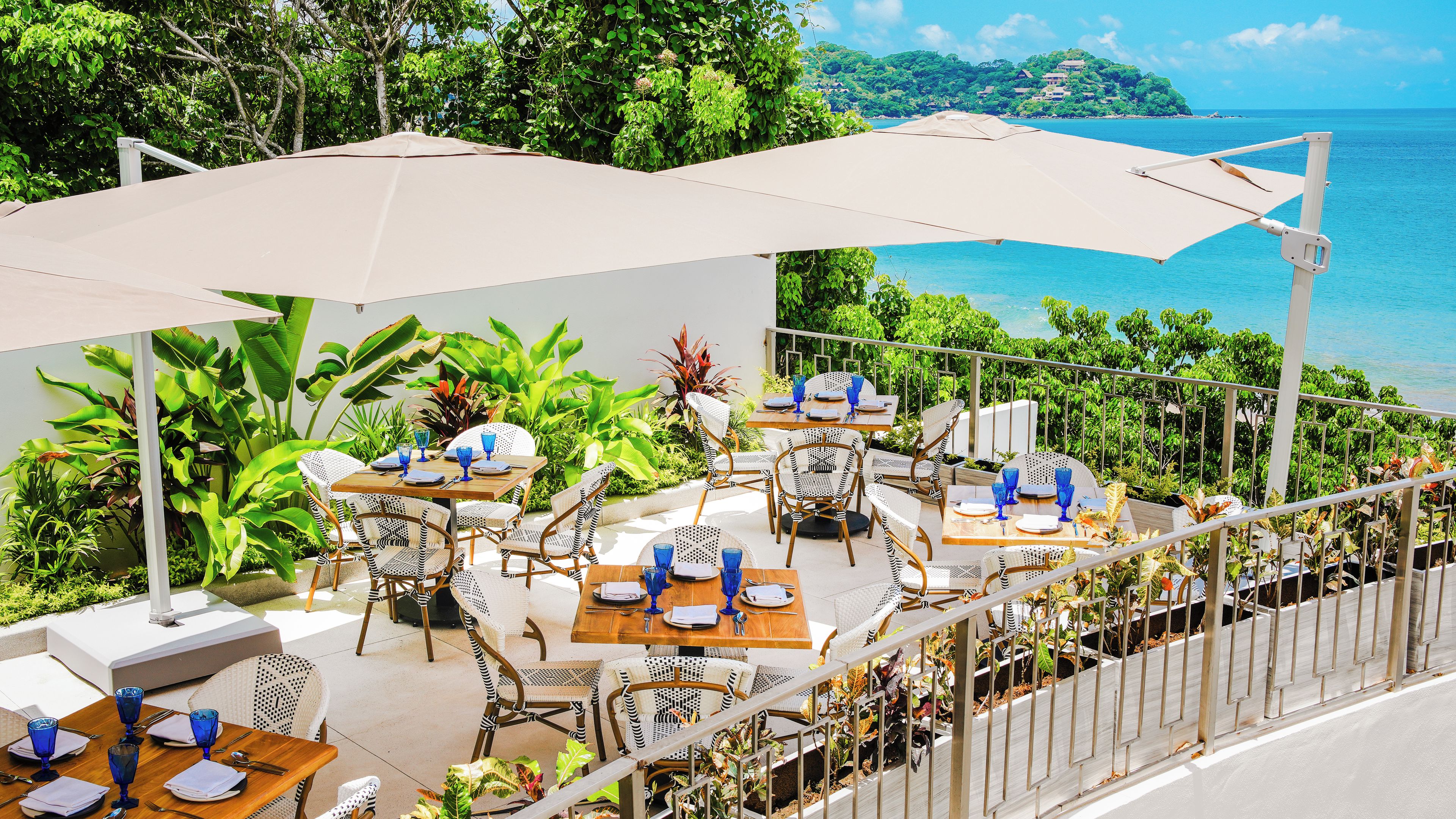 An outdoor dining area with tables and chairs, surrounded by greenery and overlooking a body of water.