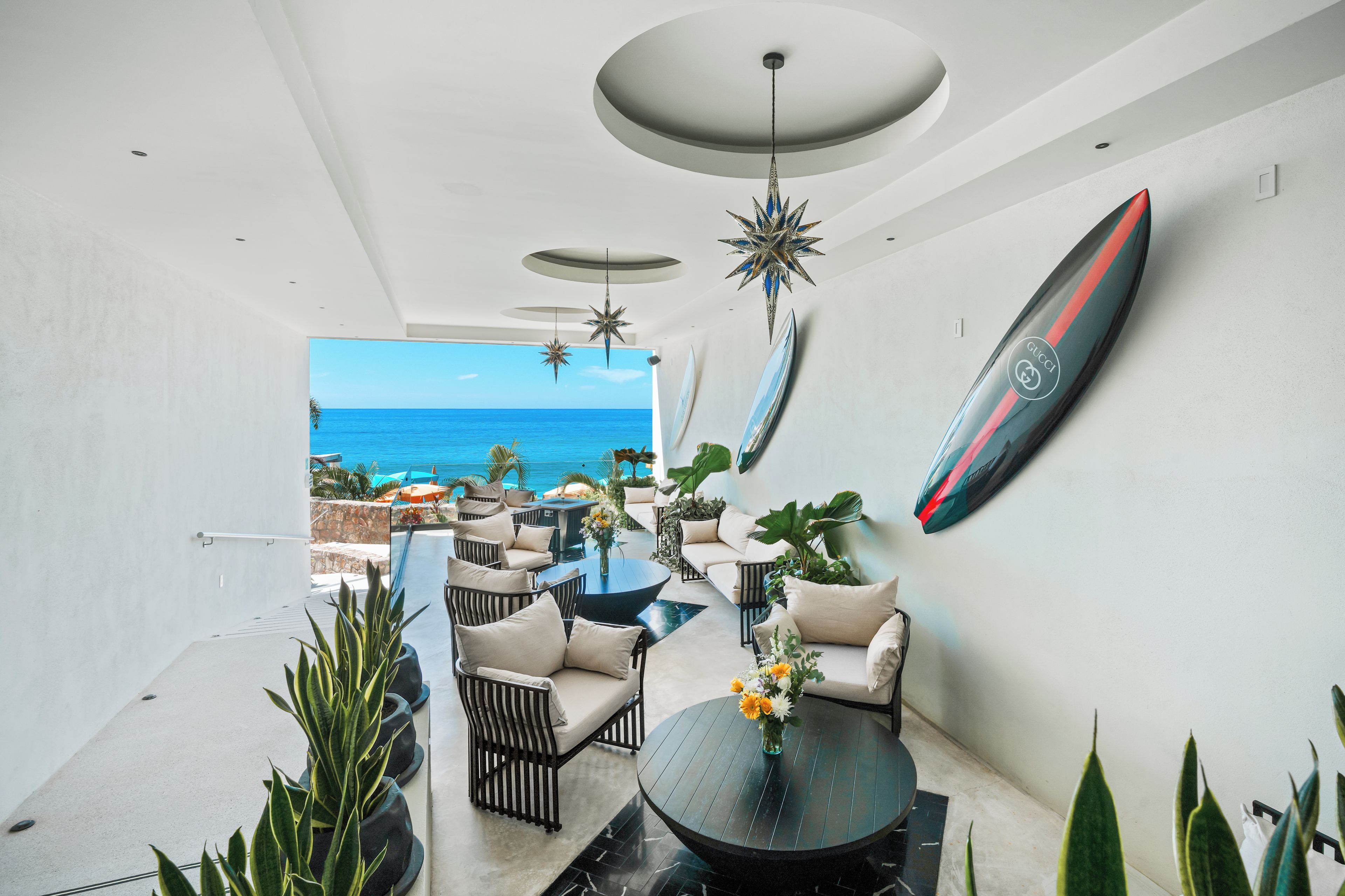 Un interior moderno con vistas al océano, que presenta un comedor con mesas y sillas redondas, una decoración con una tabla de surf y luces colgantes con forma de estrella.