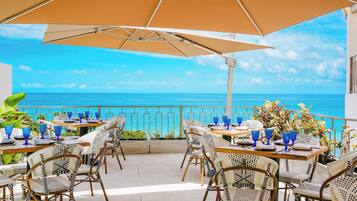 Un área de comedor al aire libre con mesas y sillas, con vista a un mar azul y transparente.