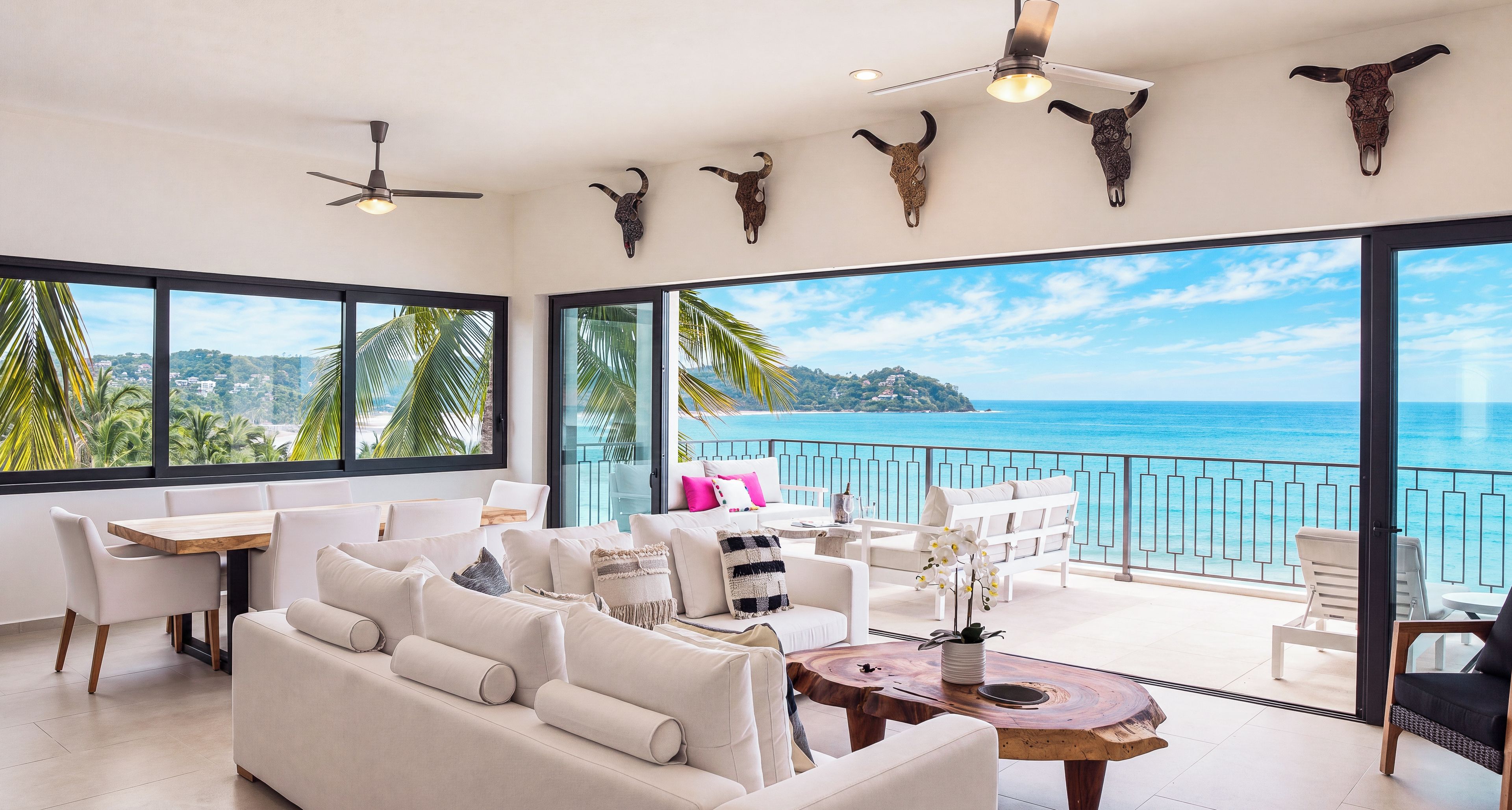 Un interior moderno con balcón y vistas al mar, que incluye un sofá blanco, una mesa de comedor y ventiladores de techo.