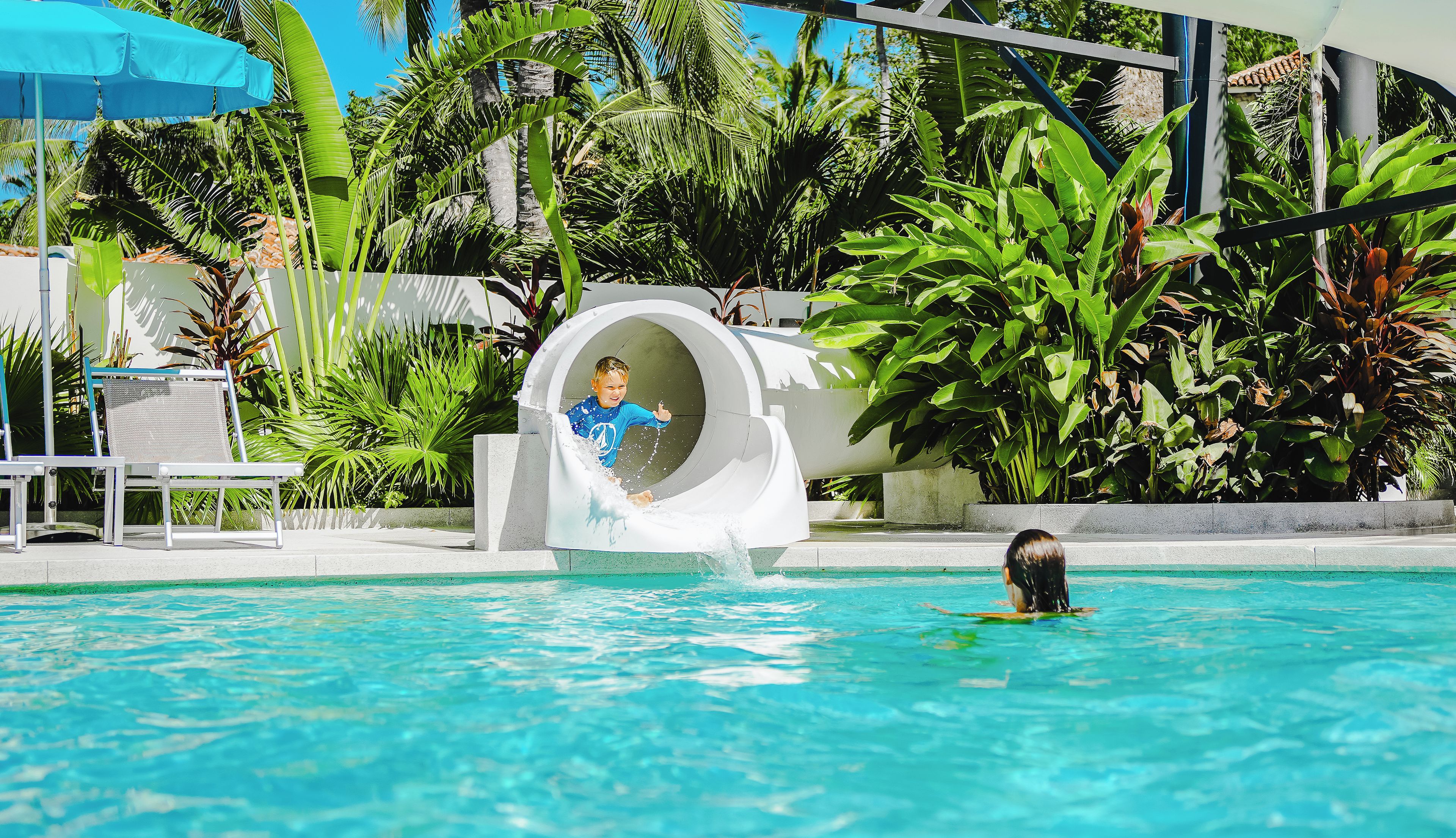 Un tobogán de agua junto a una piscina, con sillones de descanso y plantas tropicales al fondo.