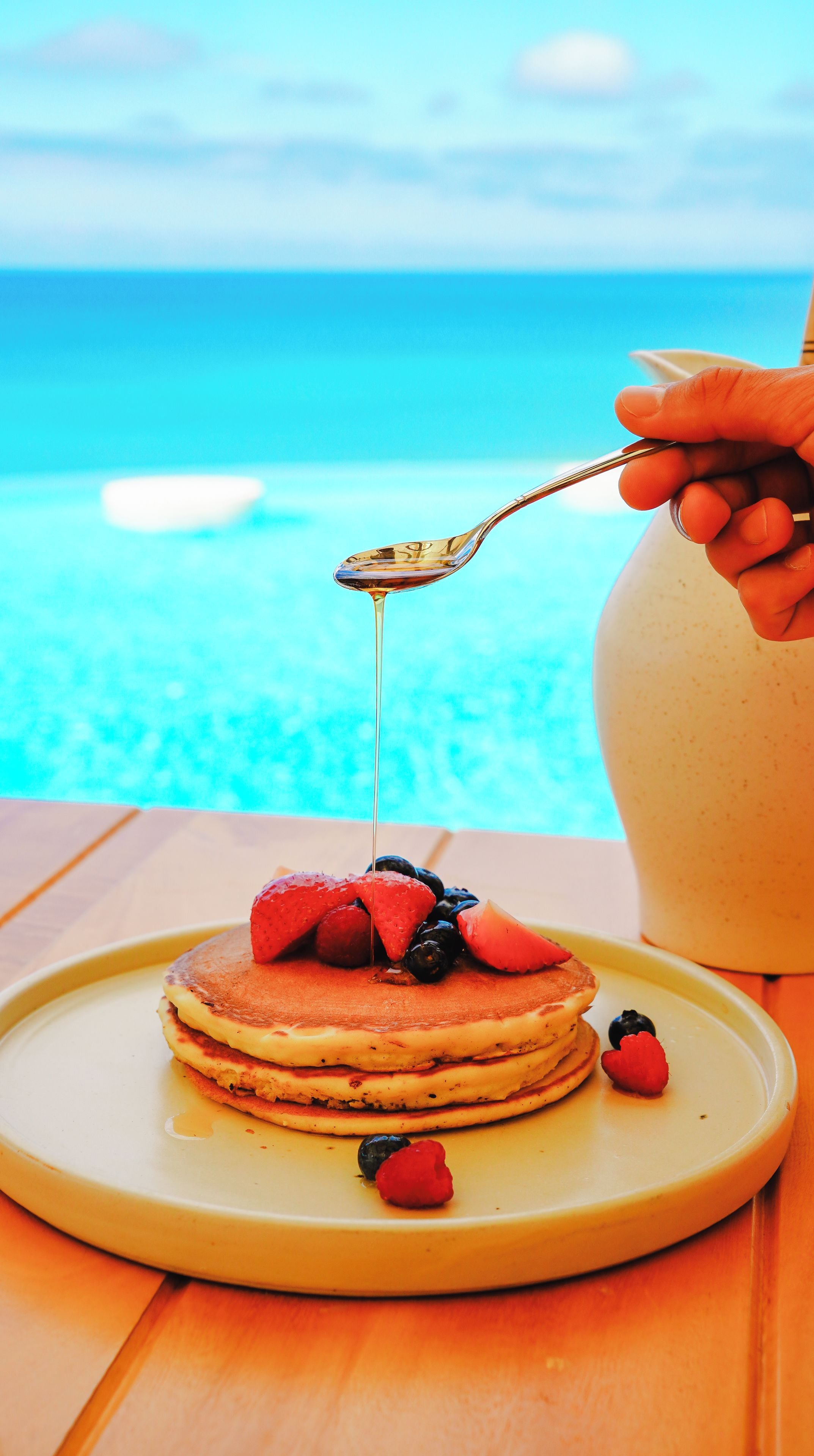 A stack of pancakes with syrup and berries on a plate, with a spoon pouring syrup, set against a backdrop of a clear blue sky and ocean.