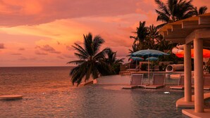 Ein Poolbereich mit Liegestühlen und Sonnenschirmen, Palmen und Blick auf das Meer bei Sonnenuntergang.