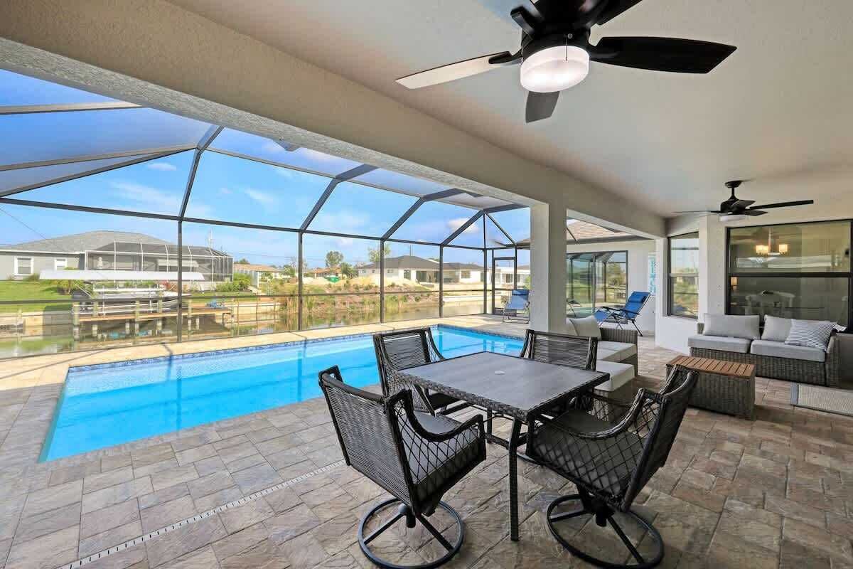 A covered outdoor swimming pool with a table and chairs, a ceiling fan, and a view of a residential area.