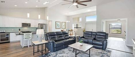 A spacious living area with a black leather sofa, a coffee table, and a dining table with chairs. There is a kitchen area with white cabinets and a microwave.