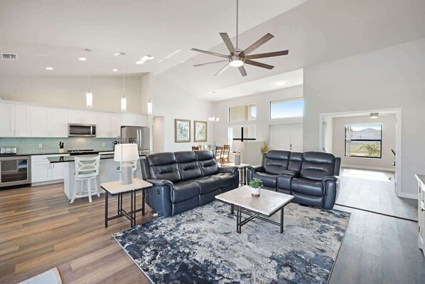 A spacious living area with a black leather sofa, a coffee table, and a dining table with chairs. There is a kitchen area with white cabinets and a microwave.