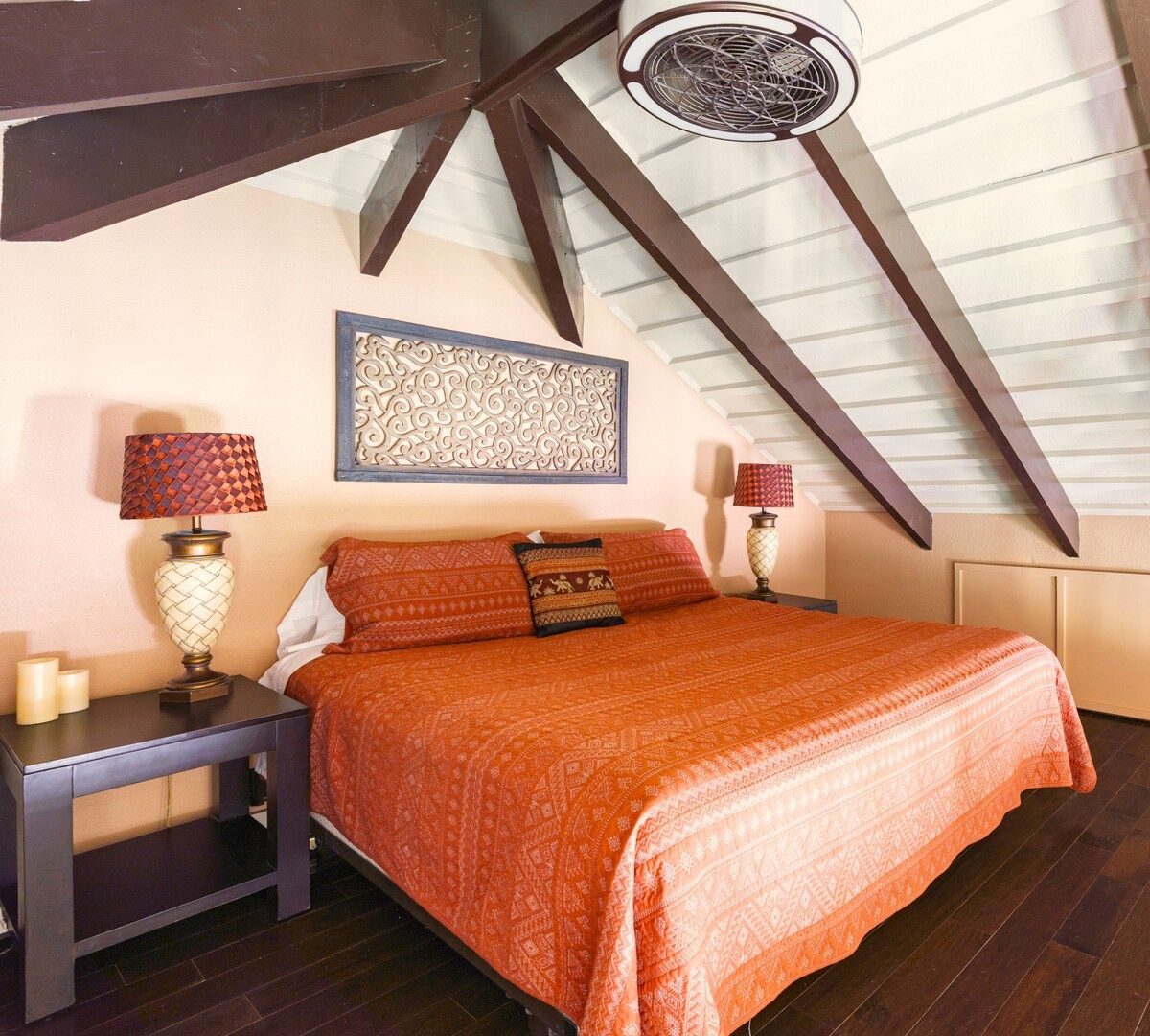 Ein Schlafzimmer mit schräg angesetzter Decke, einem Bett mit gemusterten Betttextilien, zwei Nachttischen mit Lampen und einem dekorativen Wandbild.