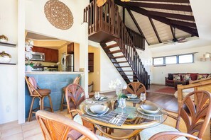 A spacious living area with a dining table set for four, surrounded by wicker chairs. A staircase with wooden railings leads to an upper level.