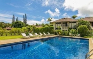A swimming pool with lounge chairs, surrounded by greenery and a building with a balcony.