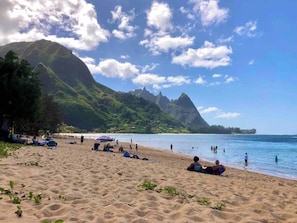 Ein Strand mit badenden und sonnenden Personen, umgeben von Bergen und blauem Himmel.