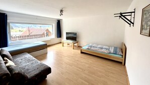 A room with a large window, a bed, a sofa, a TV, and a wooden floor. - Ferienwohnung Göss (Leoben)