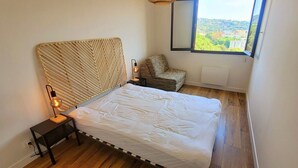 A bedroom with a bed, bedside table, lamp, chair, window, and view of the outside.