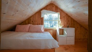 Ein gemütliches Schlafzimmer unter dem Dach mit holzverkleideter Schräge, einem großen Bett mit weißer Bettwäsche, einem kleinen weißen Nachttisch und einem Fenster mit Blick auf Bäume.