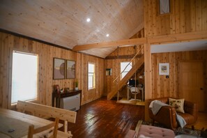 L’intérieur d’un chalet en bois comprend un coin repas, un salon avec un canapé et un fauteuil, un escalier et une petite cuisine.