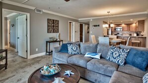 A living room with a grey sofa, a coffee table with decorative items, and a view into the kitchen area. - Beach House 304D - Gulf Front Condo (Miramar Beach)