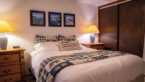 Una cama bien tendida con una manta a cuadros, dos lámparas de noche, una mesita de madera y una pared con tres cuadros enmarcados.