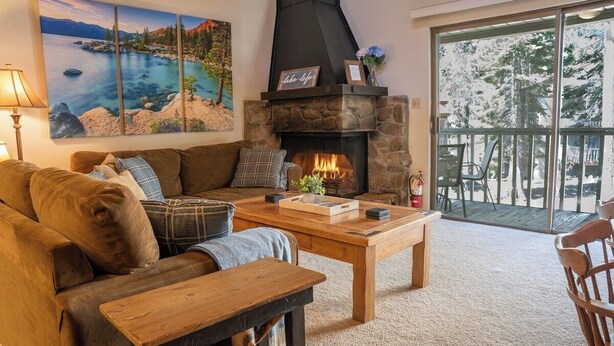 Una sala de estar acogedora con chimenea de piedra, un cuadro grande de un lago y un balcón con mobiliario exterior.