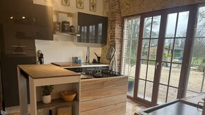 A modern kitchen with a central island, wooden cabinets, and a large window providing a view of the outdoors. - Une Longere de la Puisaye en Fond Dimpasse Avec Parc Apaisant (La Ferté-Loupière)