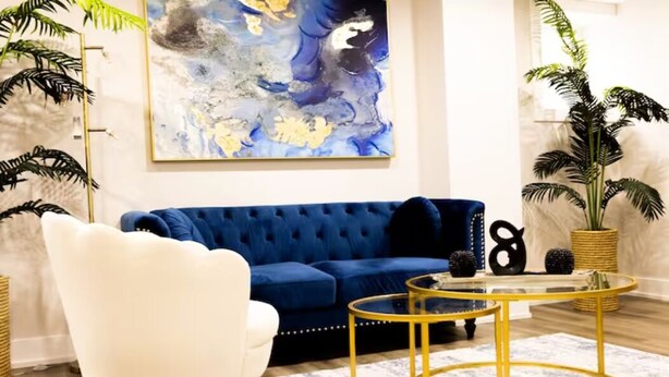A modern living room with a blue sofa, white armchair, and a glass coffee table with gold legs.