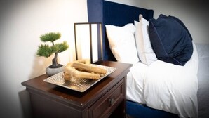 A neatly made bed with white and dark blue bedding, a small bedside table with a bonsai tree and a tray of bread rolls, and a framed mirror.
