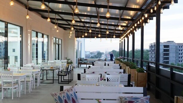 An outdoor seating area with white tables and chairs, hanging light bulbs, and large windows offering city views. - Reikartz Abis Tashkent (Tashkent)