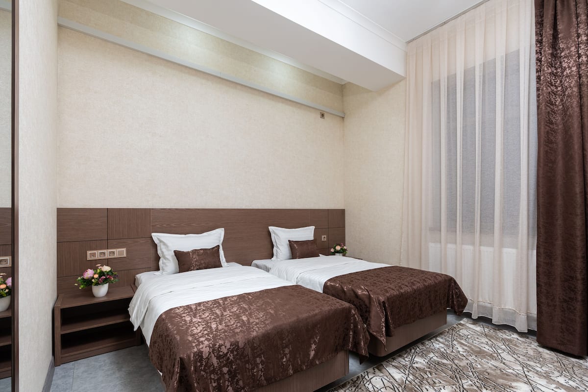 A hotel room with two beds, a wooden headboard, a small bedside table with a vase of flowers, and a large window with sheer curtains.