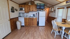 A compact kitchen with wooden cabinets, a microwave, a stove, and a refrigerator. There is a dining table with white chairs and a small window with blinds. - The Lakeview at Northside Marina & Resort (chico)