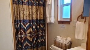 Uma casa de banho com teto de madeira, cortina de chuveiro estampada, sanita, lavatório com espelho e janela.
