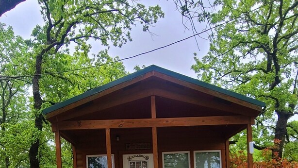 Uma pequena cabana de madeira com varanda, um letreiro de boas-vindas e uma porta.
