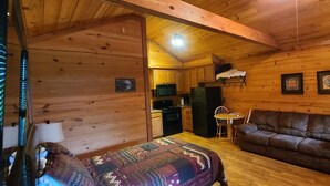Ein gemütliches Cabin-Zimmer mit Holzvertäfelung, einem Bett mit gemustertem Überwurf, einem braunen Ledersofa, einem kleinen Esstisch mit Stühlen und einer Küchenzeile inklusive Kühlschrank und Herd.