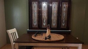 Uma sala de jantar com mesa de madeira, cadeiras brancas, um jogo americano tecido e um aparador decorativo com portas de vidro.