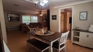 Uma sala de jantar com mesa e cadeiras de madeira, um candelabro e vista para a cozinha e a sala de estar.