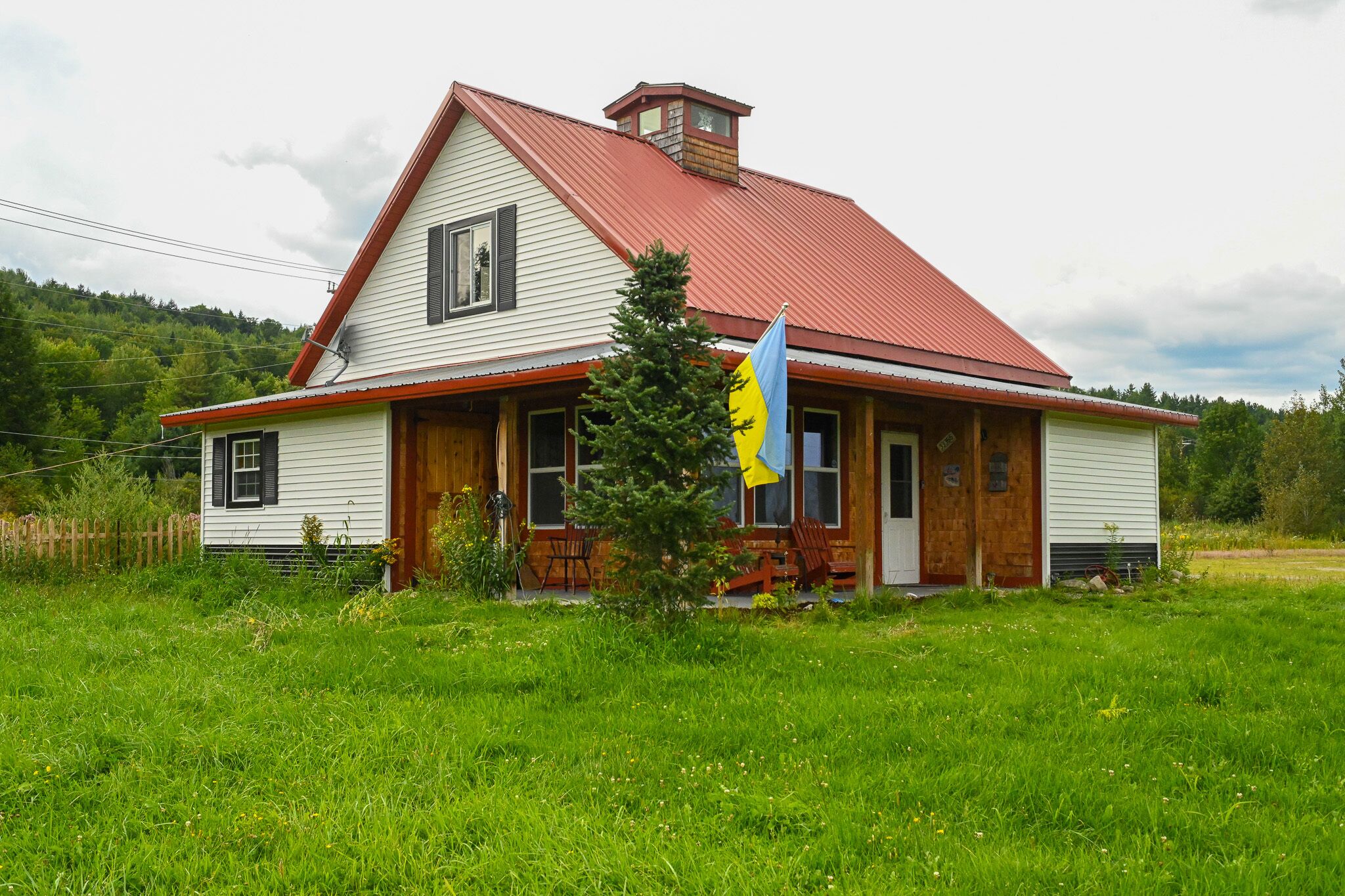 Uma casa térrea com telhado vermelho e revestimento branco, um alpendre com cadeiras de madeira e um mastro com uma bandeira.