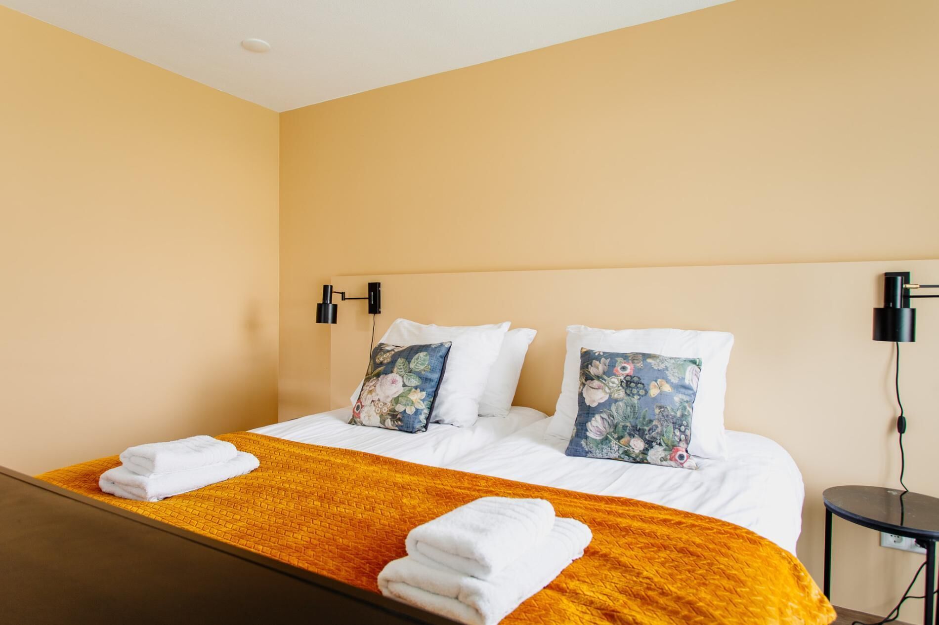 Ein Hotelzimmer mit Doppelbett, zwei Kissen mit Blumenmuster, einer gelben Tagesdecke und zwei Wandlampen.
