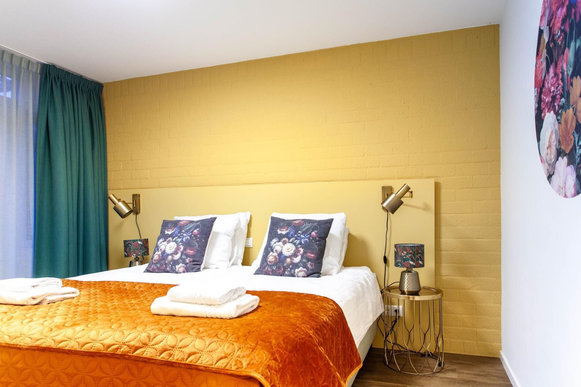 Ett hotellrum med en stor säng, två vägglampor, ett runt sidobord och ett blommönstrat konstverk på väggen.