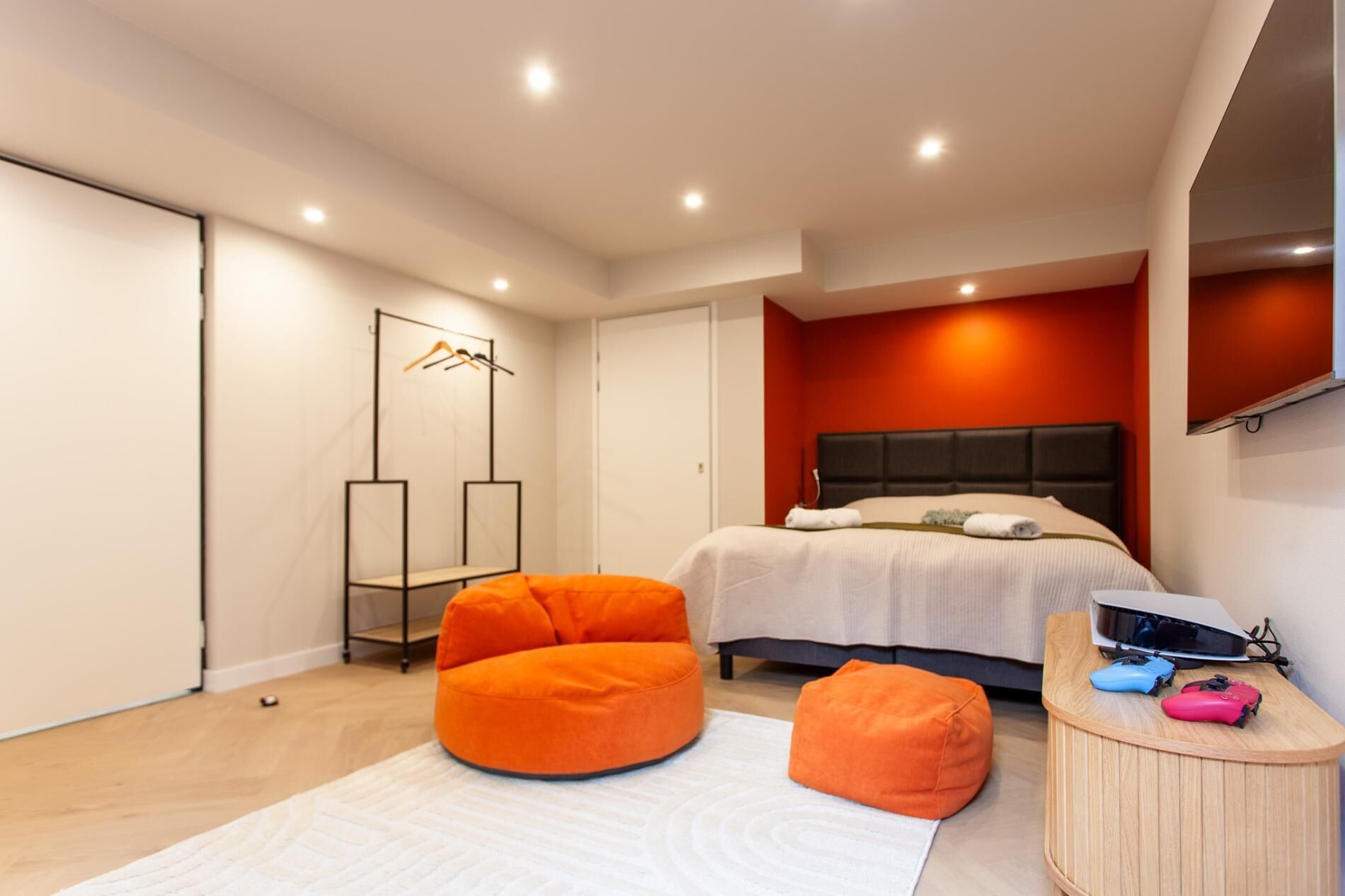Een moderne slaapkamer met een bed, oranje poefs, een houten nachtkastje met een laptop en hoofdtelefoon, en een spiegel met kleding.