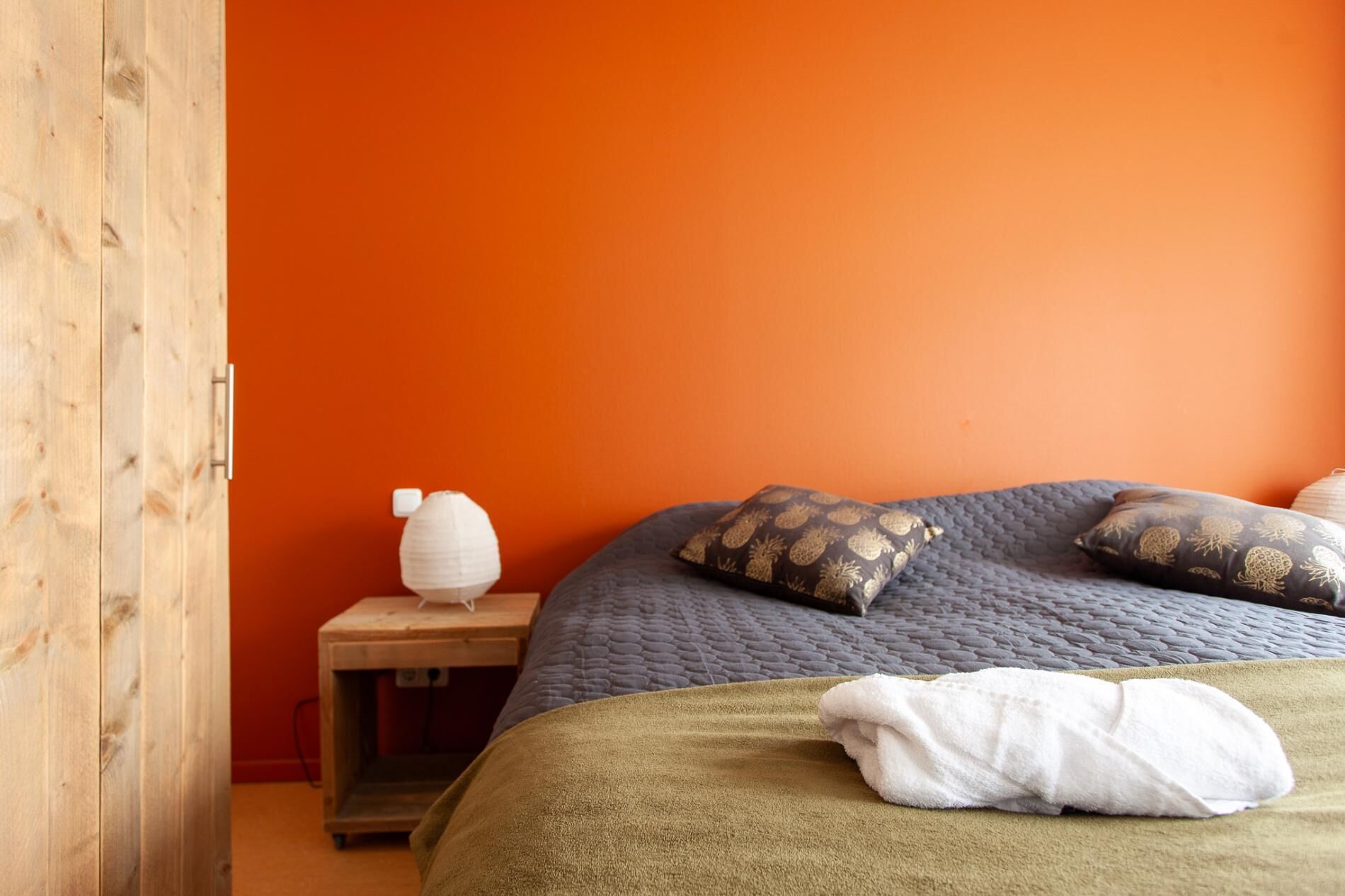 A bedroom with an orange wall, a wooden wardrobe, a bedside table with a lamp, a bed with a green blanket and patterned pillows, and a white towel on the bed.