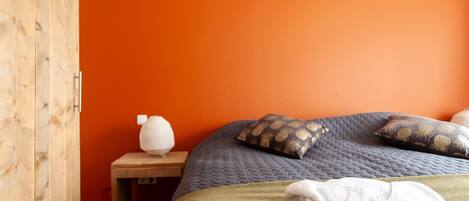 A bedroom with an orange wall, a wooden wardrobe, a bedside table with a lamp, a bed with a green blanket and patterned pillows, and a white towel on the bed.