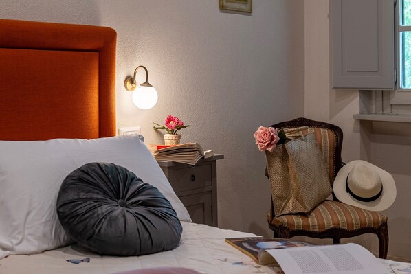 Een netjes opgemaakt bed met een kussen, een nachtkastje met een lamp, een stoel met een hoed, en een raam met gordijnen.