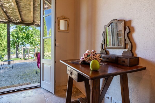 Un escritorio de madera con un espejo, un florero con flores y una puerta que da a un espacio exterior con un banco y árboles.