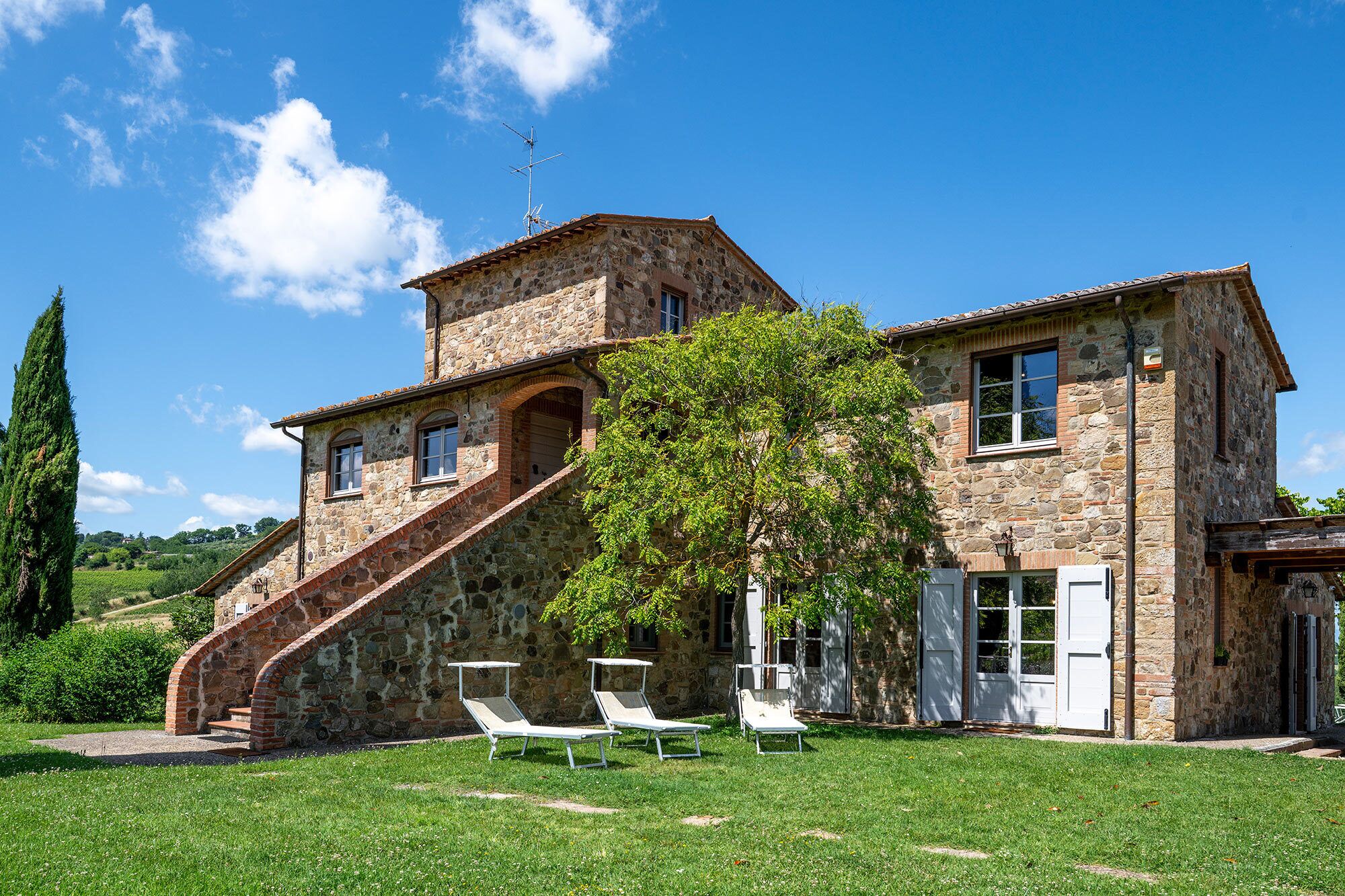 A stone building with a staircase leading to a second floor, a tree in the front yard, and three white lounge chairs.
