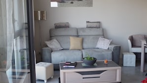 Una sala de estar moderna con un sofá gris, una mesa de centro y una pequeña mesa auxiliar. Hay una puerta de cristal que da a un balcón, y un cuadro de una escena de playa en la pared.