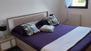 Un dormitorio ordenado con una cama de madera, ropa de cama morada, una lámpara de noche, una ventana con cortinas translúcidas y un radiador.