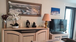 Una sala de estar moderna con comedor, que incluye un televisor de pantalla plana grande, una lámpara decorativa y un cuadro de una escena costera.