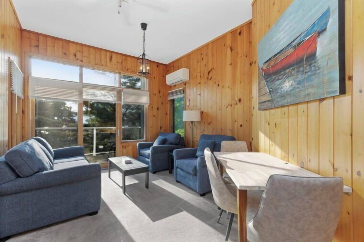 A wooden-paneled room with a sofa, two armchairs, a dining table, and a painting of a boat on the wall.