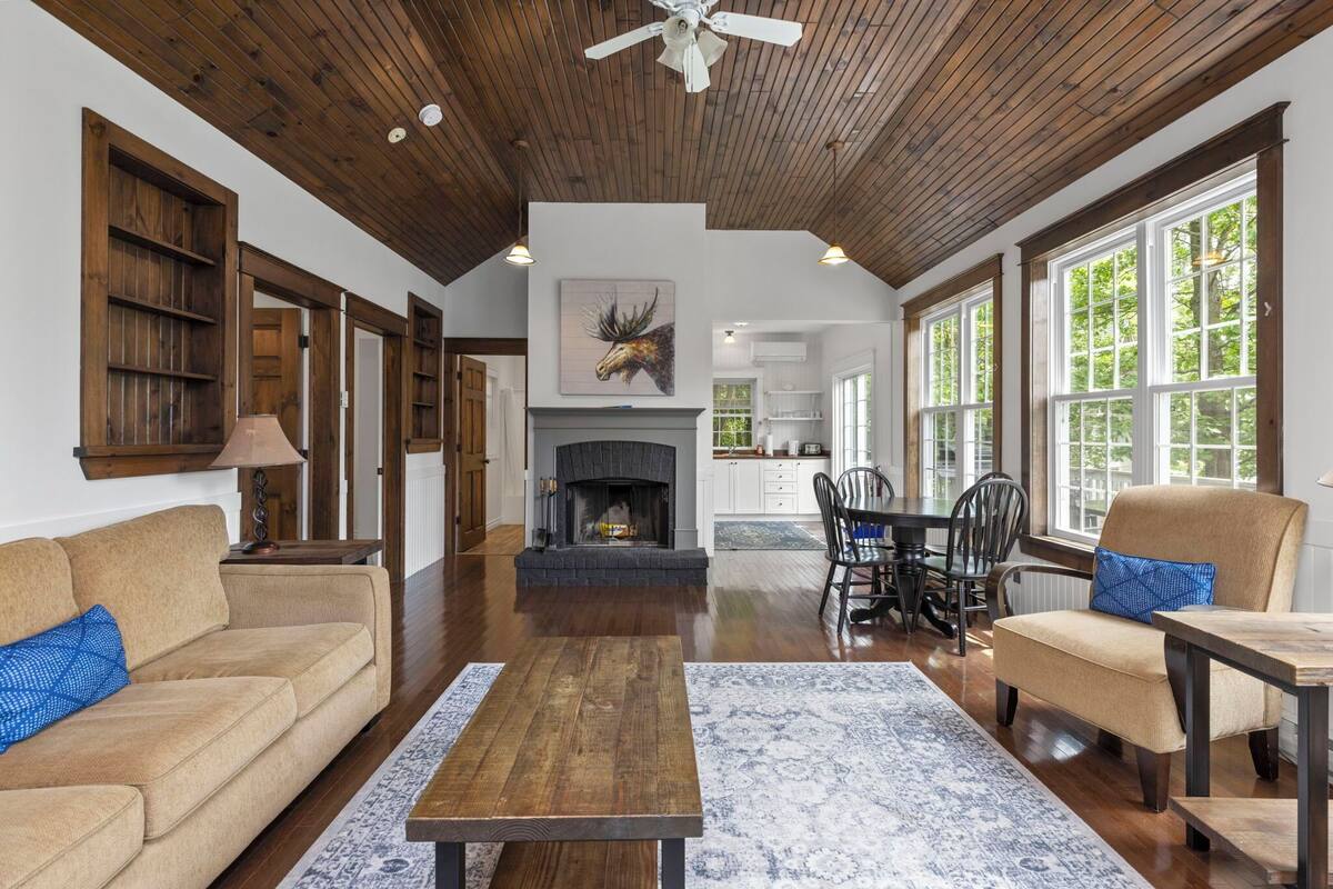 A spacious living room with a fireplace, wooden ceiling, and large windows.