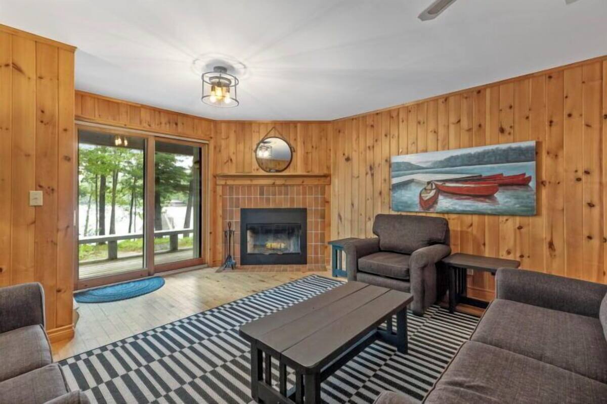 A cozy living room with wooden walls, a fireplace, a large window with a view of trees, and a painting of a lake scene.